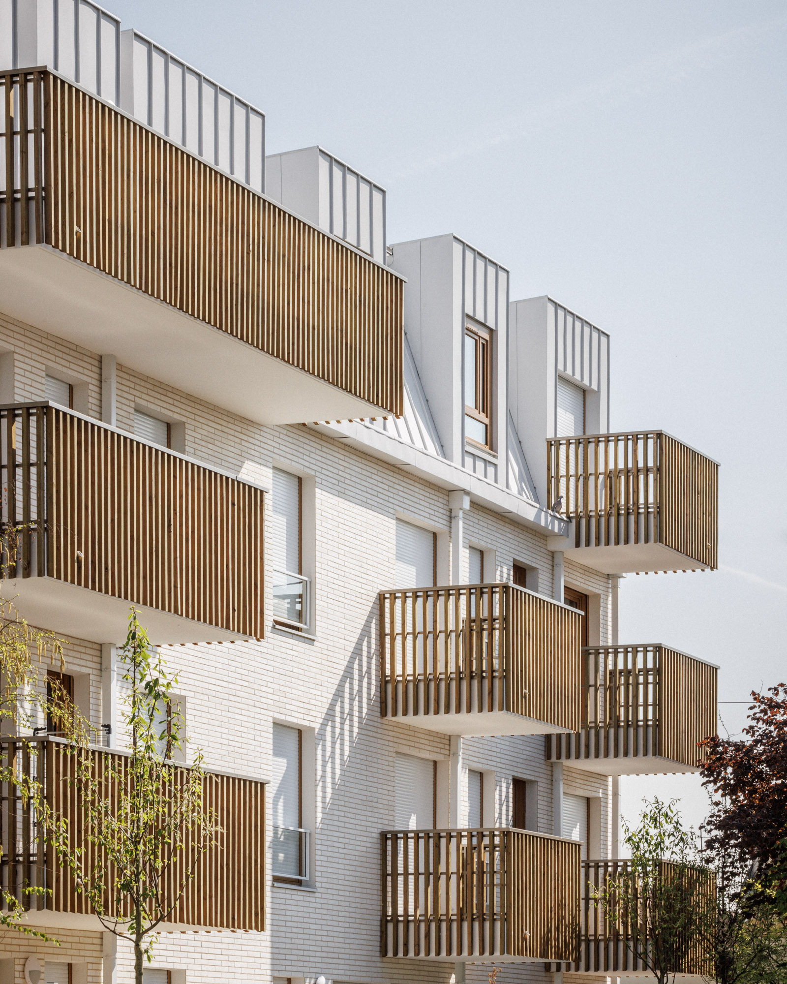 Détail des façades d'une résidence de 56 logements collectifs conçue par Cussac Architectes & Associés (CA+A) à Bondy (Seine-Saint-Denis), avec balcons en bois, façades en briques blanches et toitures en zinc à joints debout.